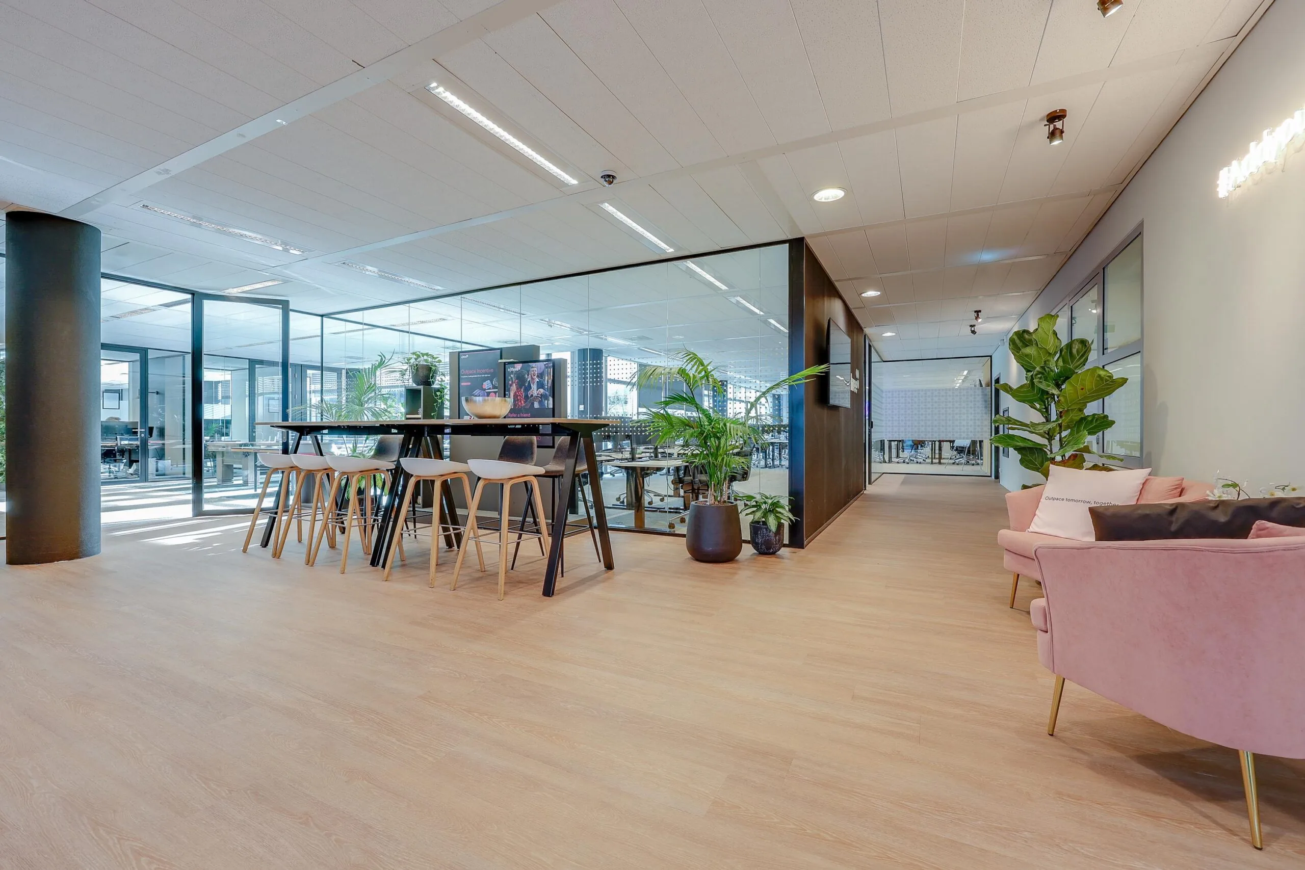 Een moderne kantoorruimte met een hoge tafel en stoelen, glazen wanden en roze zitgelegenheid.