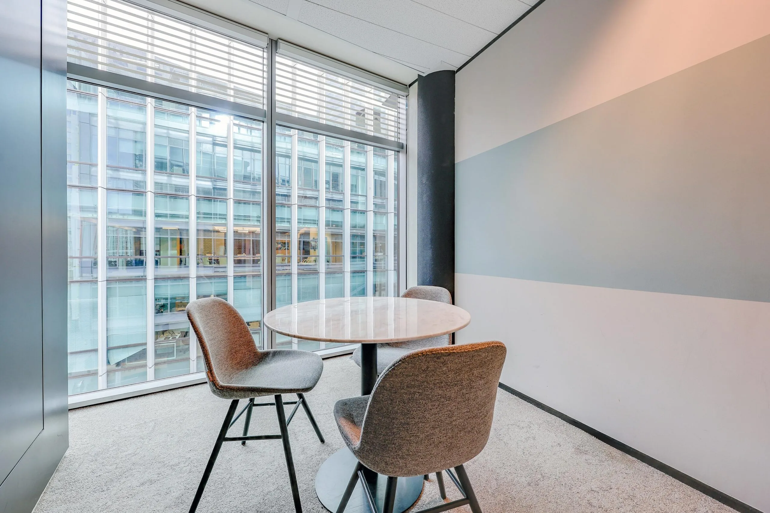 Een kleine vergaderruimte met een ronde tafel en drie stoelen, gelegen bij een raam met uitzicht op een modern kantoorgebouw.