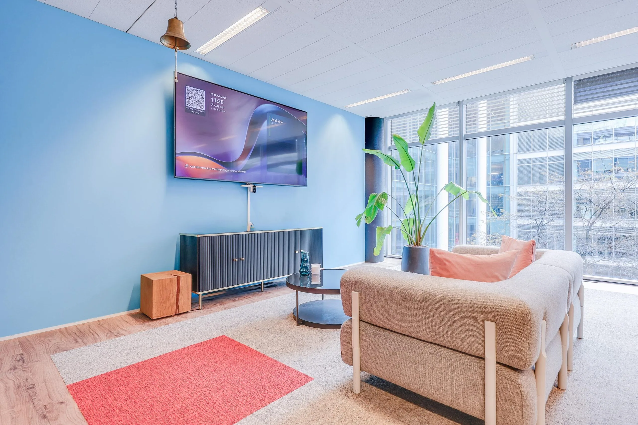 Een moderne kantoorruimte met een blauwe muur, grote ramen, een bank, een plant en een televisiescherm.