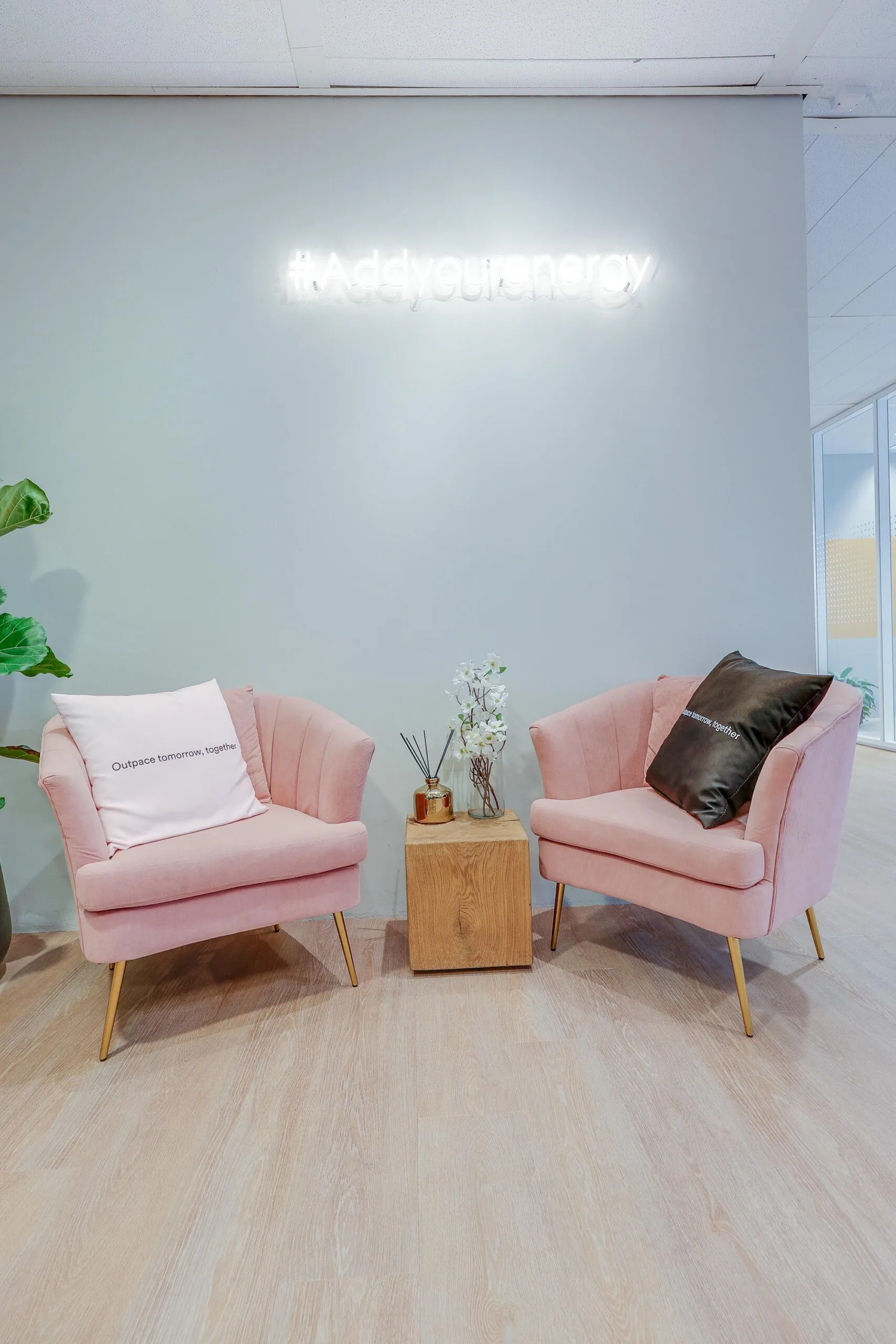 Twee roze stoelen met kussens en een bijzettafel met decoratie onder een neonbord met de tekst "#Addyourenergy".