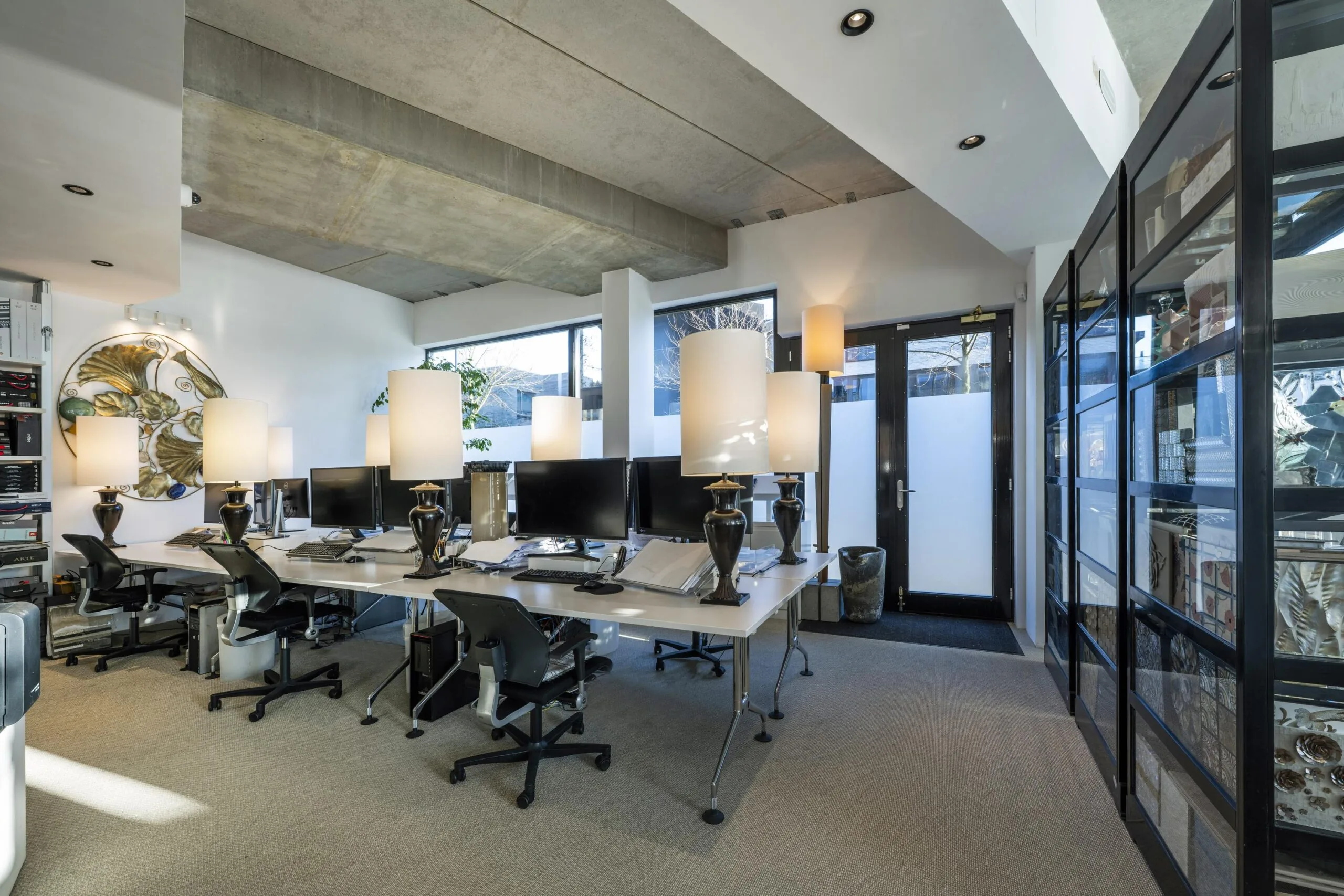 Een modern kantoor met meerdere bureaus, computers en grote tafellampen, met veel natuurlijk licht dat door ramen binnenkomt.