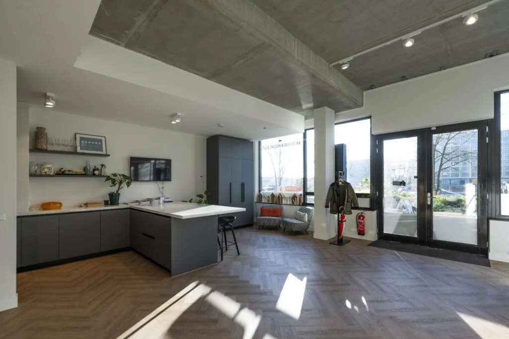 Moderne keuken-inrichting met grijze kasten en houten vloer onder een betonnen plafond met grote ramen en een zithoek.