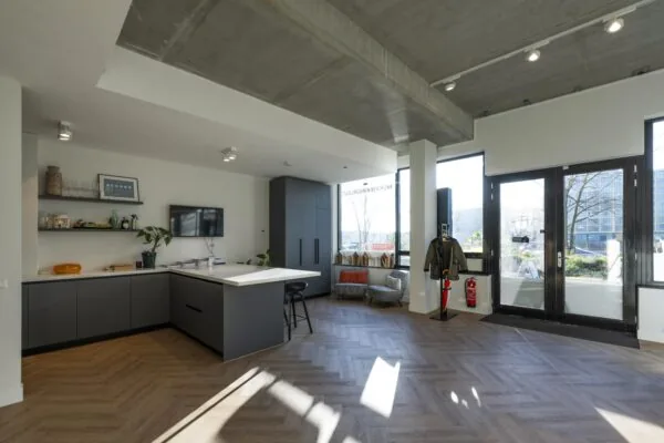 Moderne keuken-inrichting met grijze kasten en houten vloer onder een betonnen plafond met grote ramen en een zithoek.