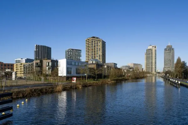 Modern stedelijk landschap langs een kanaal met hoogbouw in de verte onder een heldere blauwe lucht aan de Lindenhoevestraat.