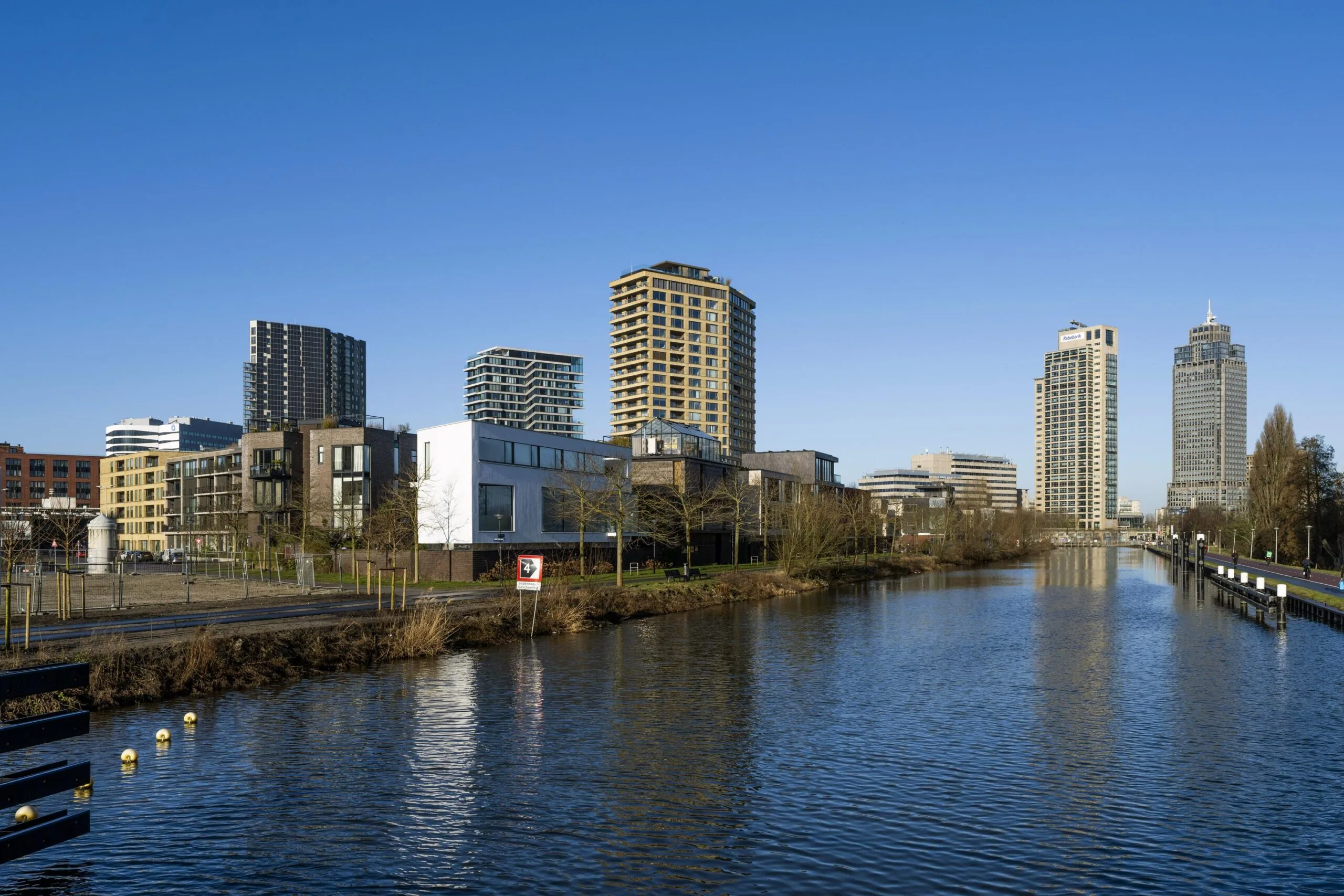 Modern stedelijk landschap langs een kanaal met hoogbouw in de verte onder een heldere blauwe lucht aan de Lindenhoevestraat.