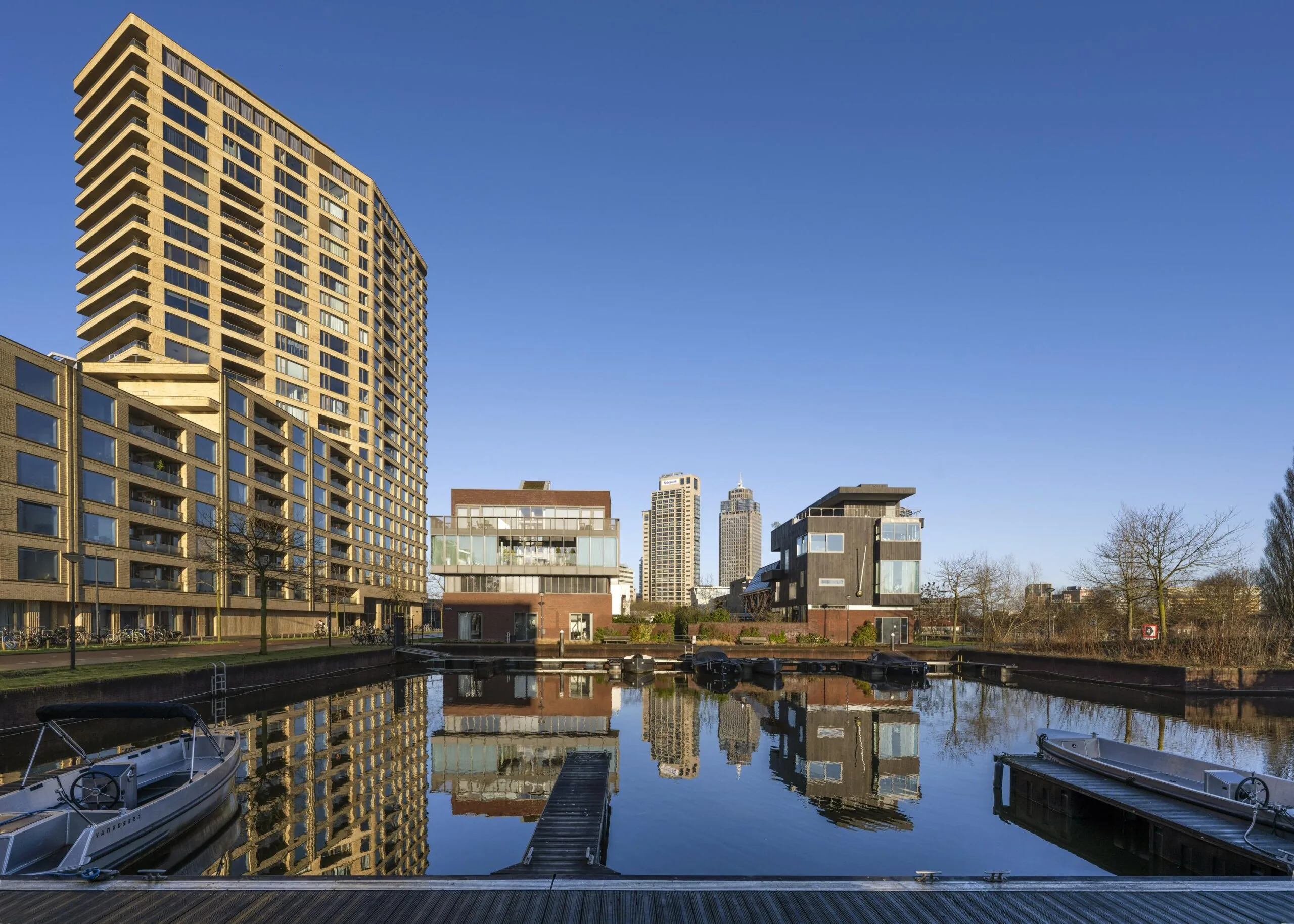 Een moderne stedelijke omgeving met hoge gebouwen die reflecteren in een kanaal, onder een heldere blauwe lucht.