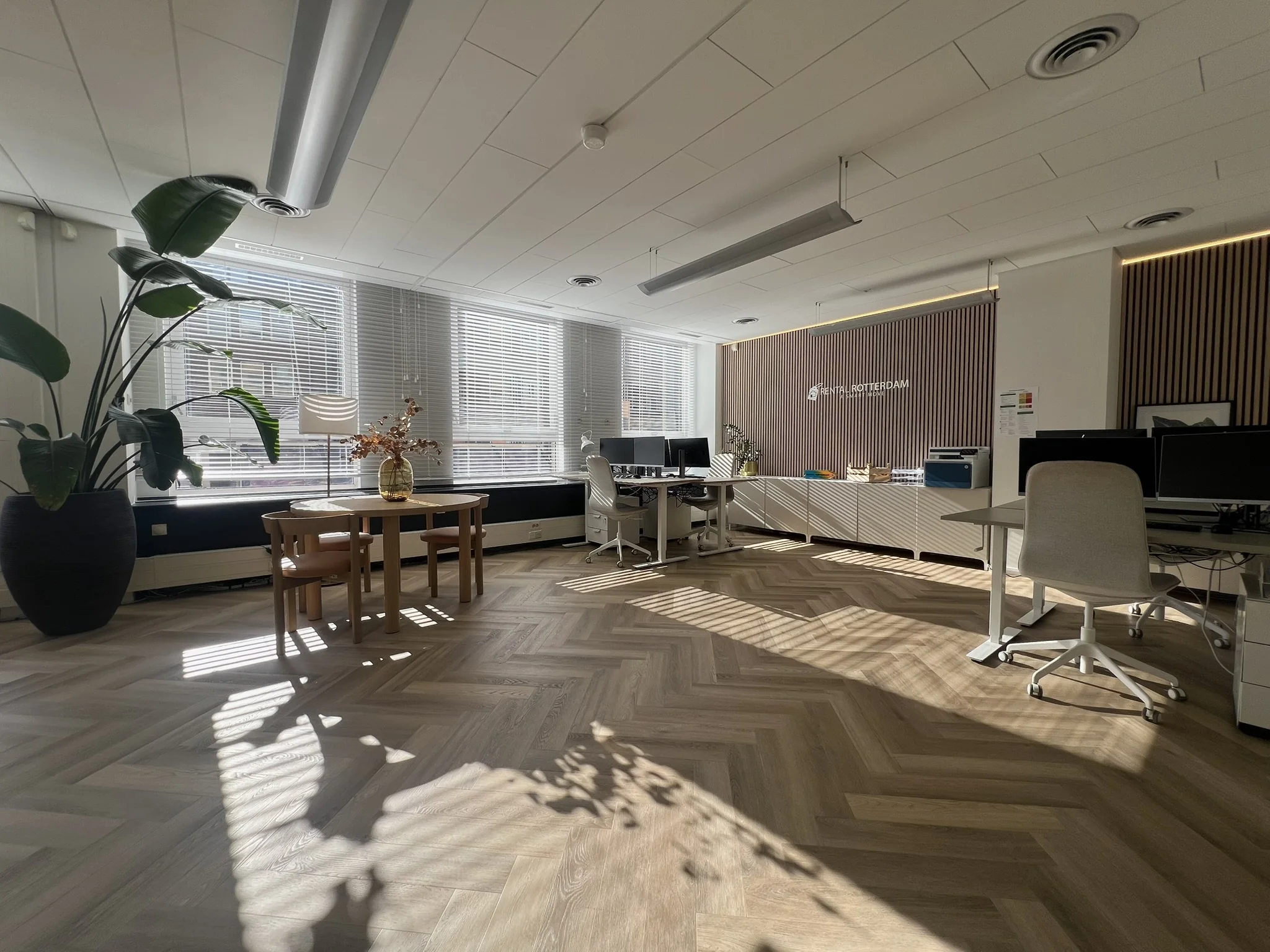 Een zonnig kantoor met houten vloer, planten en bureaus met computers.