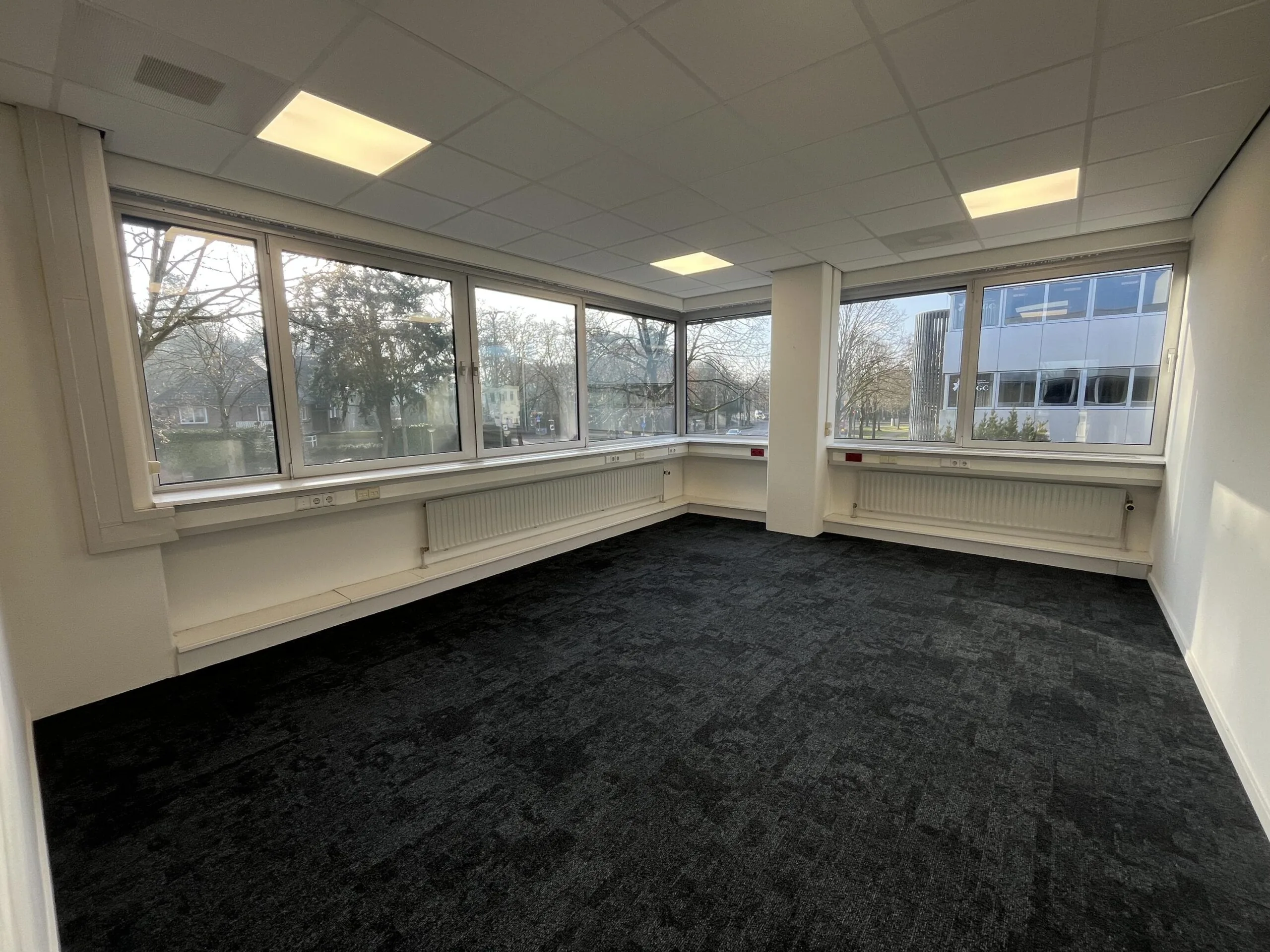 Een lege kantoorruimte met grote ramen die uitzicht bieden op bomen en een modern gebouw, gelegen aan de Huis ter Heideweg.