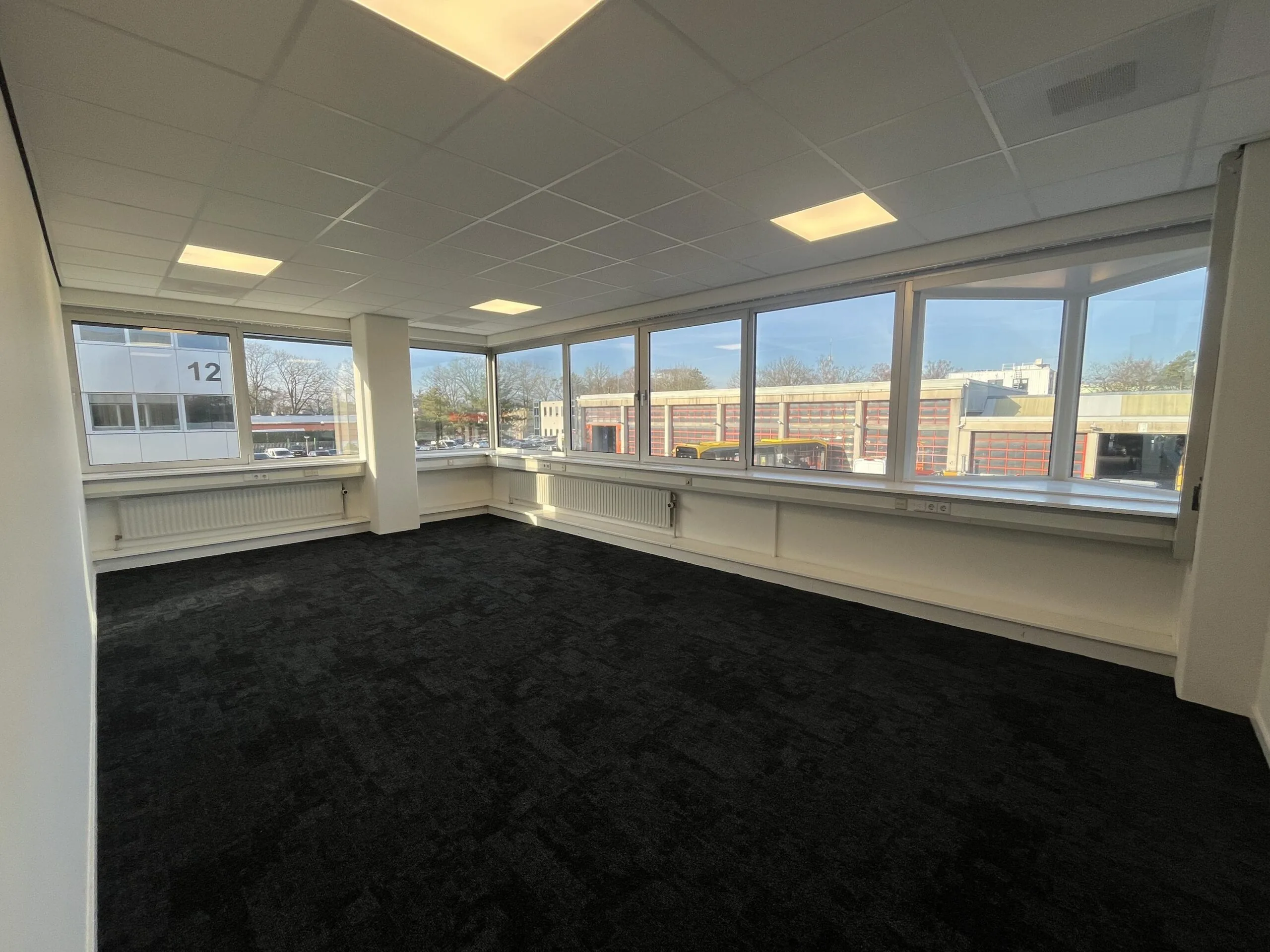 Een leeg kantoor met grote ramen aan de Huis ter Heideweg, met uitzicht op een industrieel gebouw.