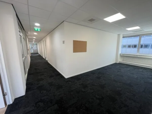 Een moderne kantoorhal met donkere vloerbedekking, tl-verlichting, en een prikbord aan de muur.