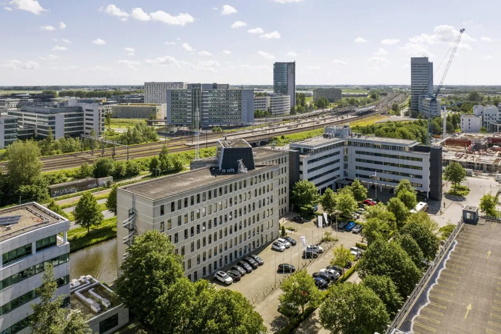 Luchtfoto van kantoorgebouwen en parkeerplaatsen aan de Neptunusstraat, met spoorlijnen en hoogbouw op de achtergrond.