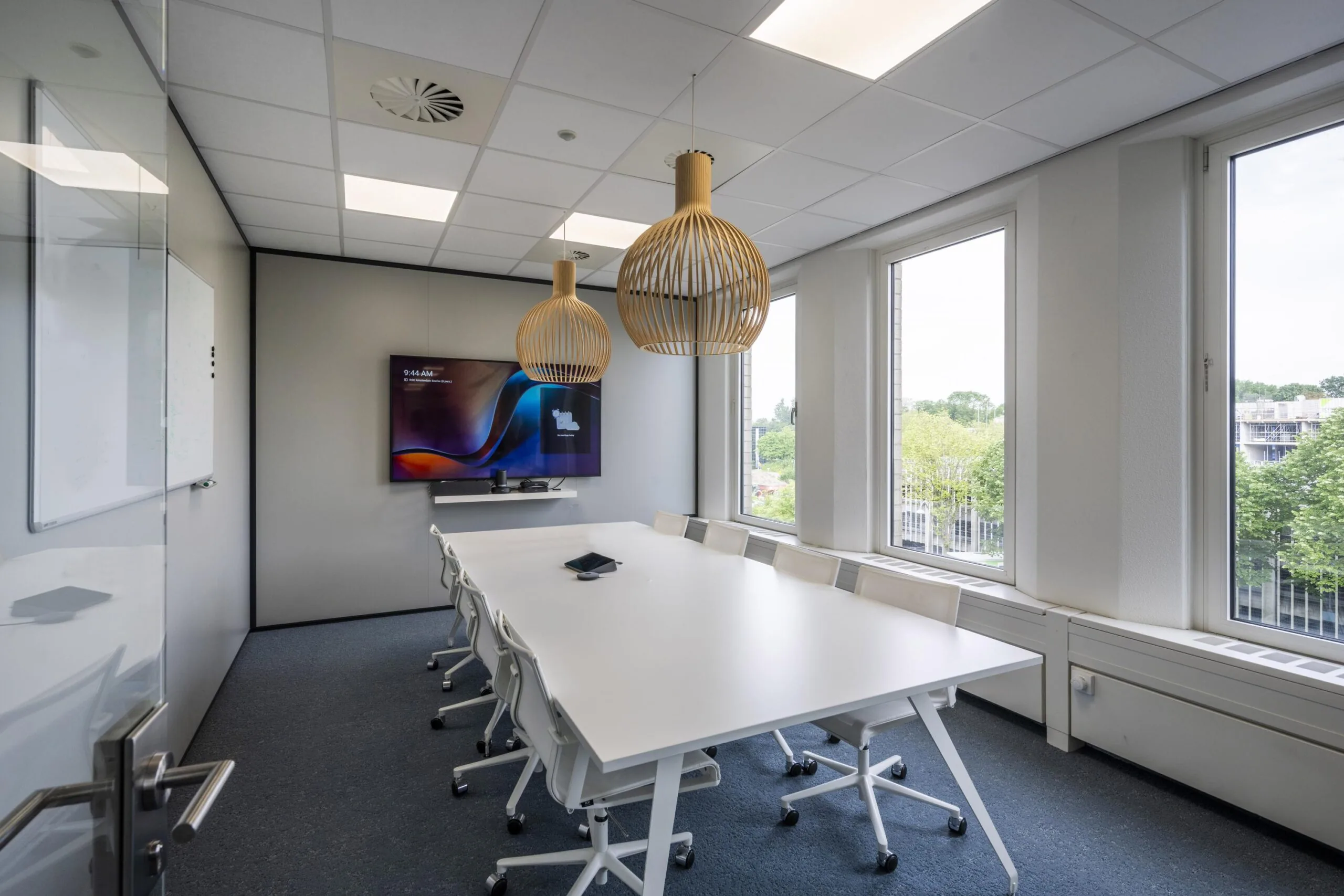 Vergaderruimte met een lange witte tafel, stoelen, hanglampen en ramen met uitzicht op buiten.
