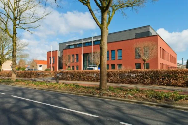Een modern rood bakstenen kantoorgebouw aan de Groenekanseweg, gefotografeerd vanaf de straat met bomen en een heg op de voorgrond.