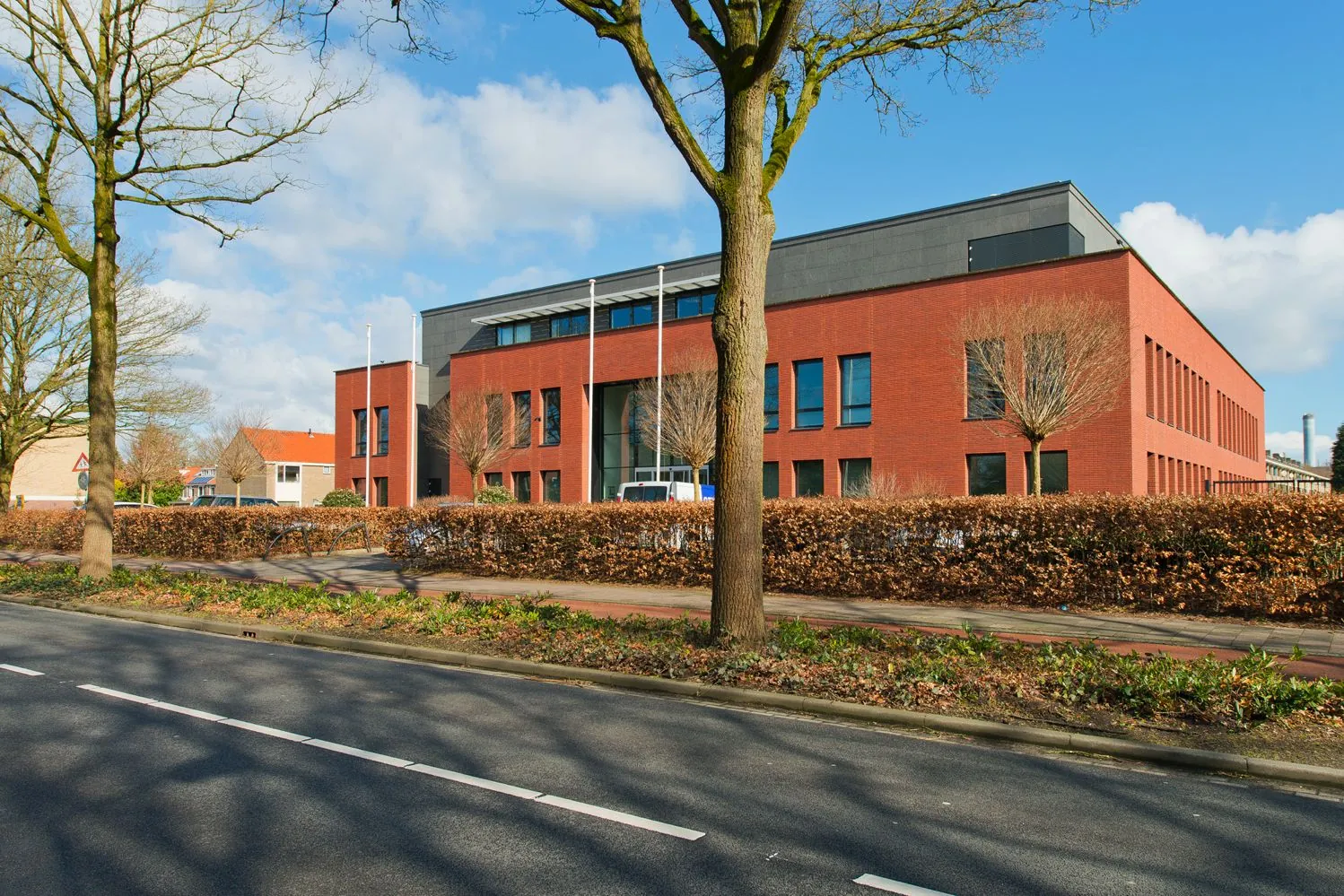 Een modern rood bakstenen kantoorgebouw aan de Groenekanseweg, gefotografeerd vanaf de straat met bomen en een heg op de voorgrond.
