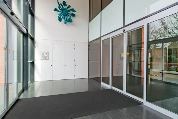Ingang van een gebouw met glazen schuifdeuren en een modern kunstwerk aan de muur.
