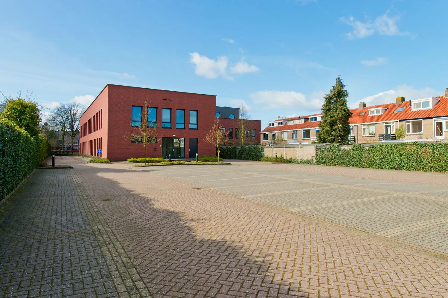 Een rood bakstenen gebouw met een lege parkeerplaats op de Groenekanseweg, omgeven door groene hagen en rijtjeshuizen.