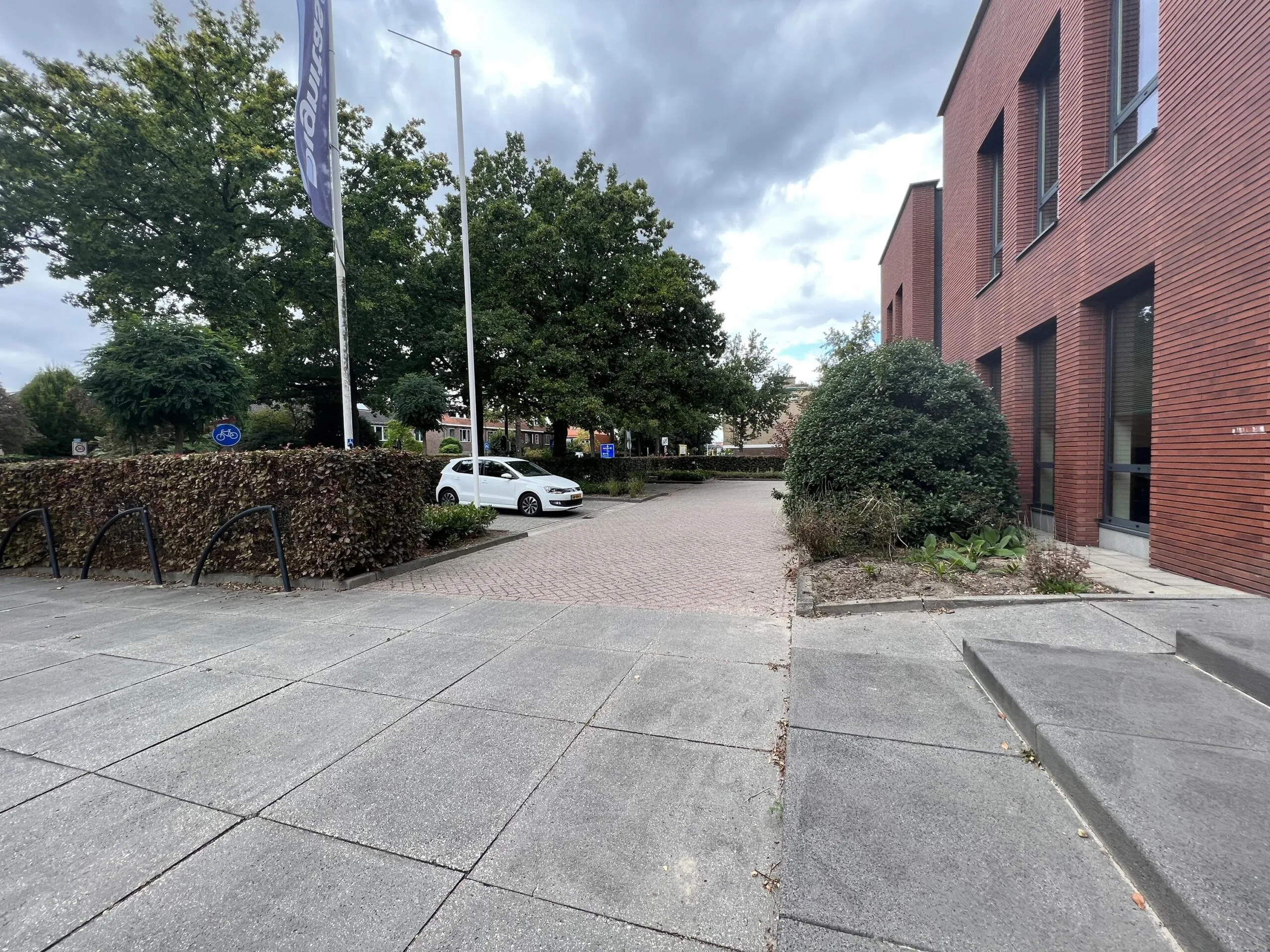 Een betegeld plein bij een modern bakstenen gebouw met een geparkeerde witte auto en groene bomen op de achtergrond.
