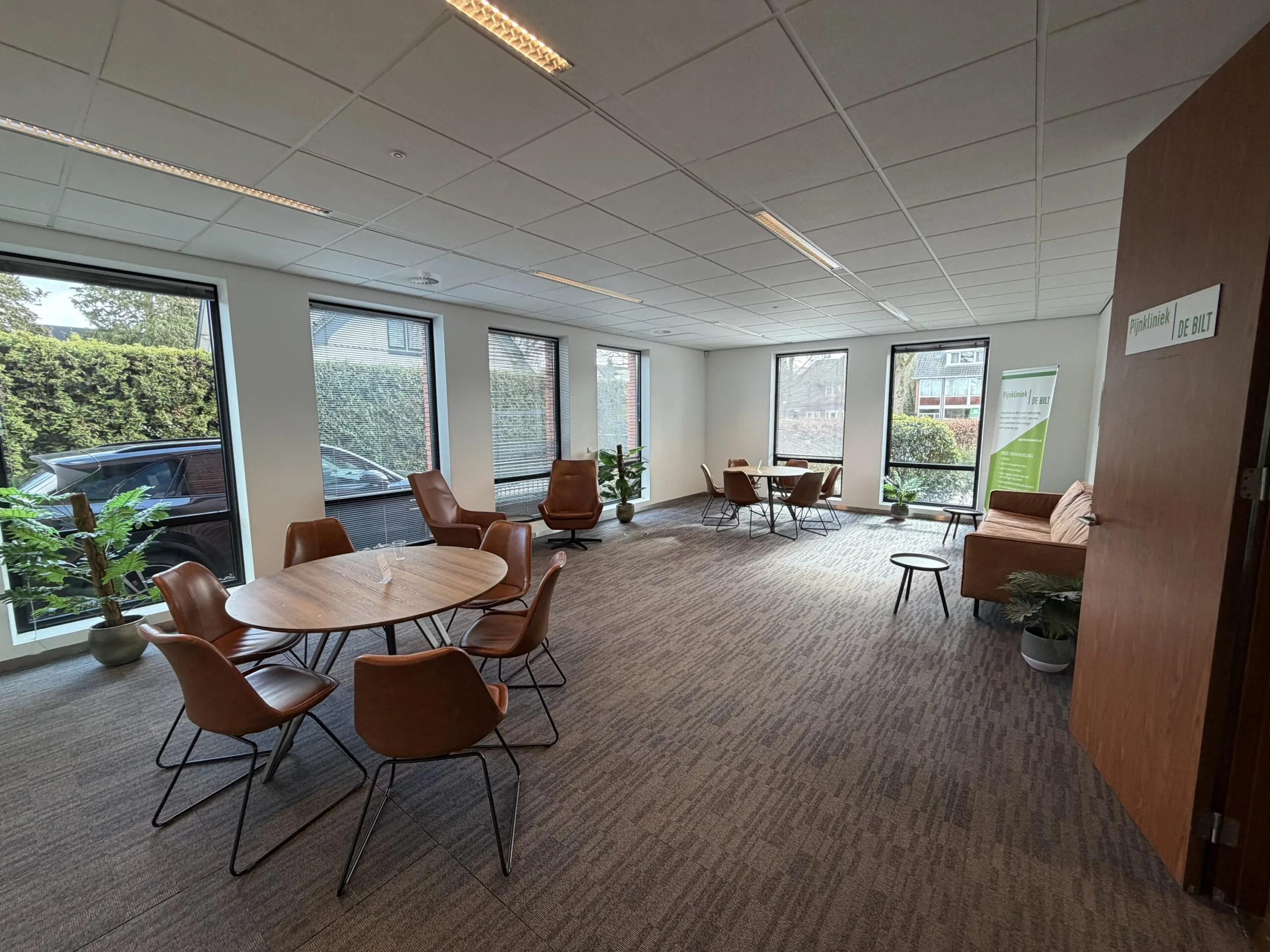 Een moderne wachtkamer met tafels en stoelen, grote ramen en enkele planten.