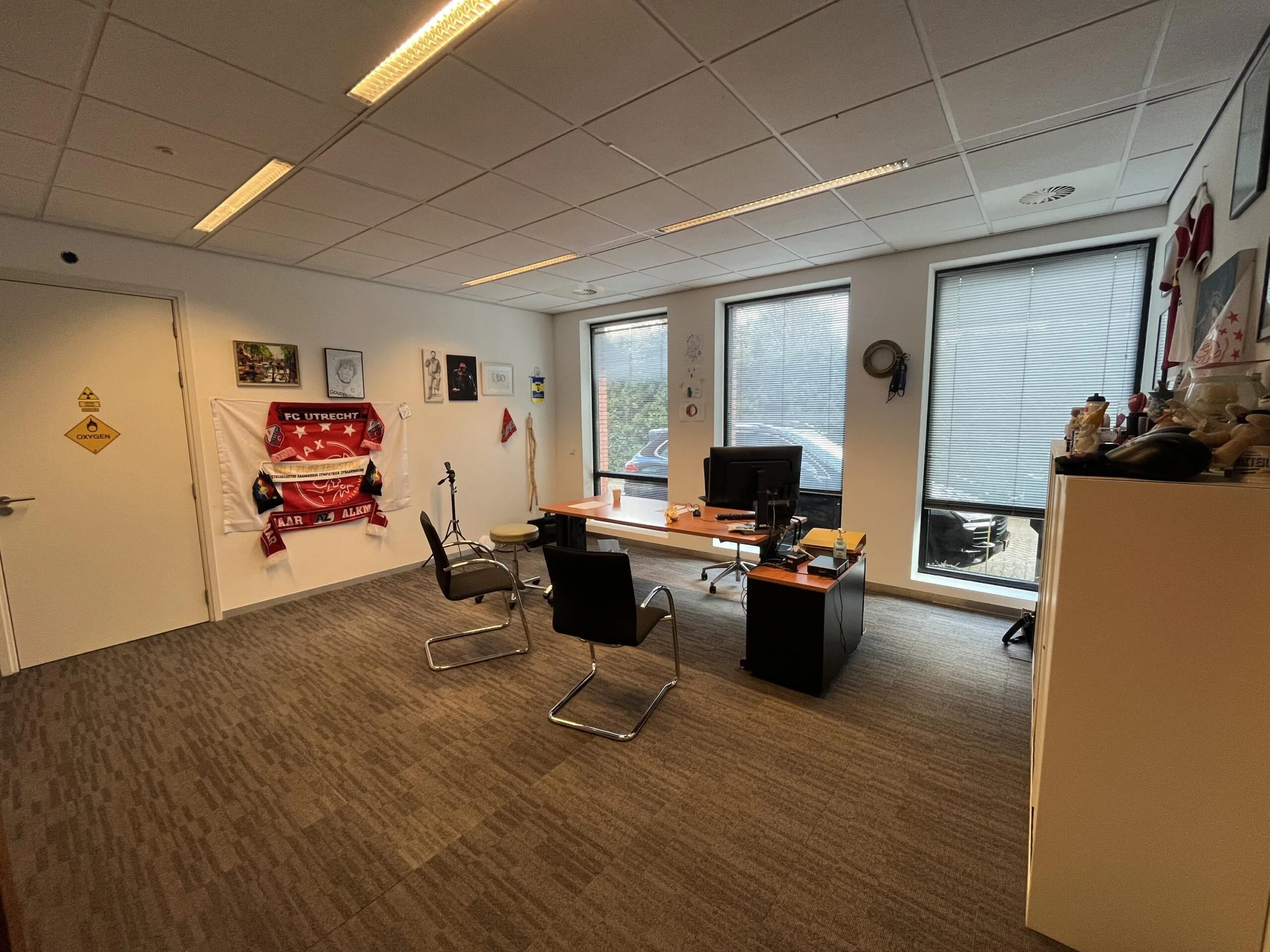 Een kantoorruimte met decoraties van FC Utrecht aan de muur, uitgerust met een bureau, stoelen en grote ramen.