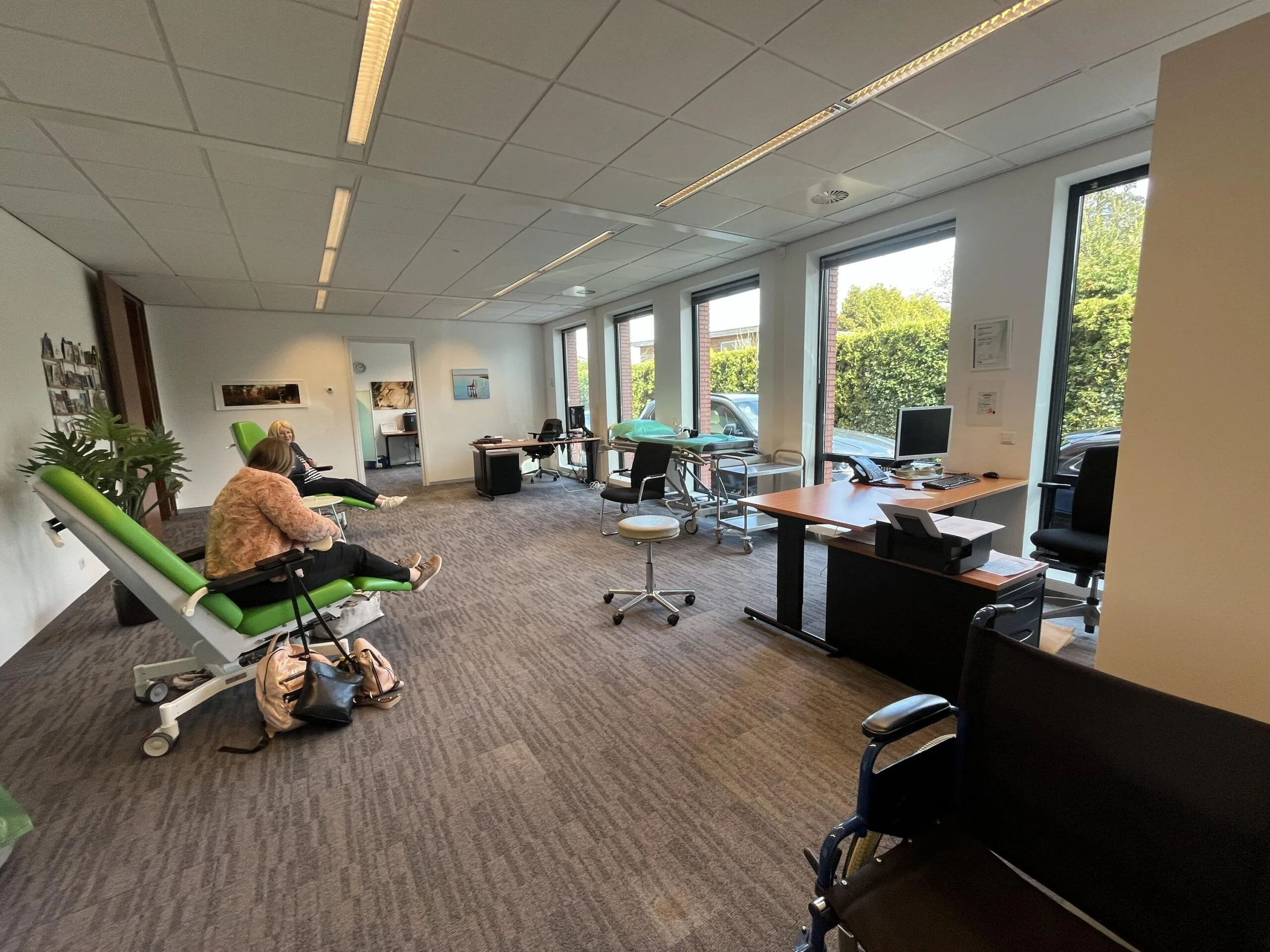Een wachtruimte met groene stoelen, mensen die zitten en een bureau bij grote ramen.