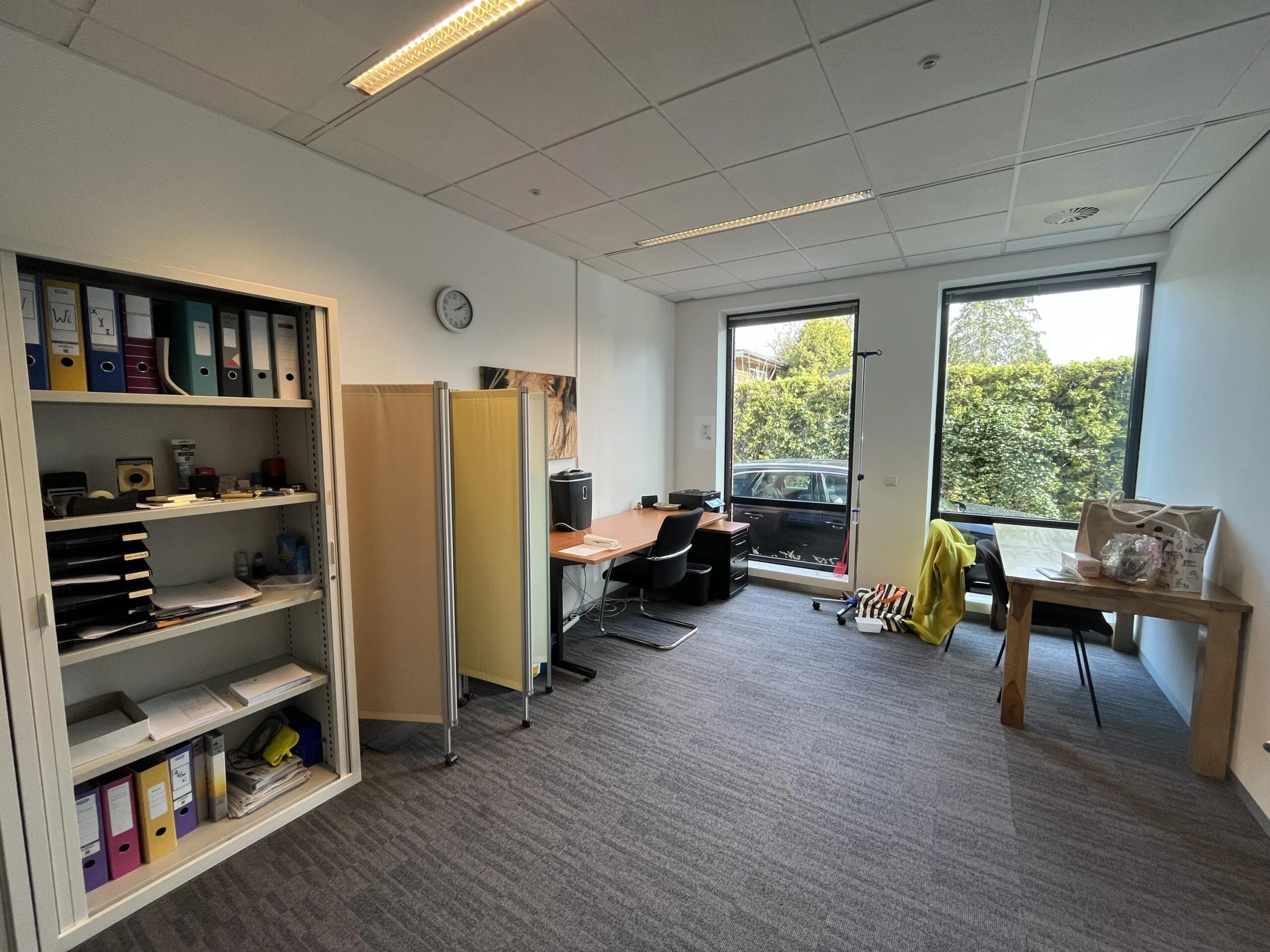 Een kantoorruimte met een kast vol ordners, een bureau bij een raam en een tafel met diverse spullen erop.