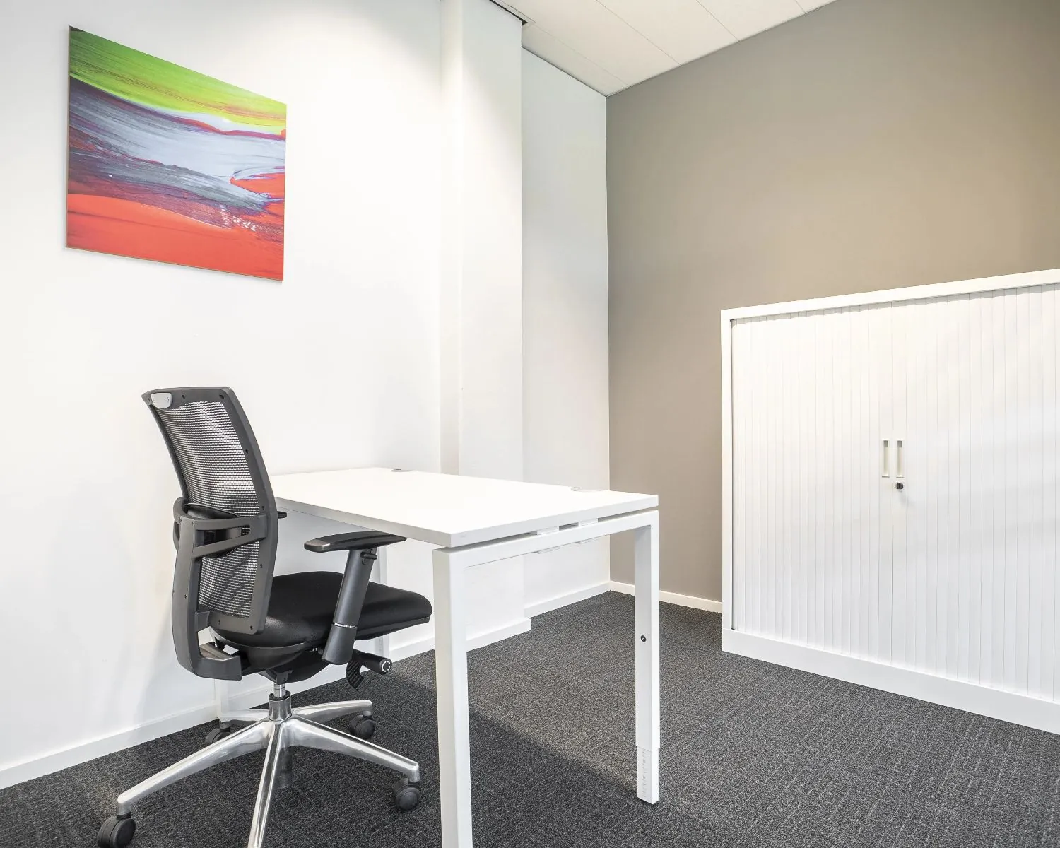 Modern en minimalistisch kantoor met een wit bureau, zwarte bureaustoel, witte opbergkast en kleurrijk abstract schilderij aan de muur.