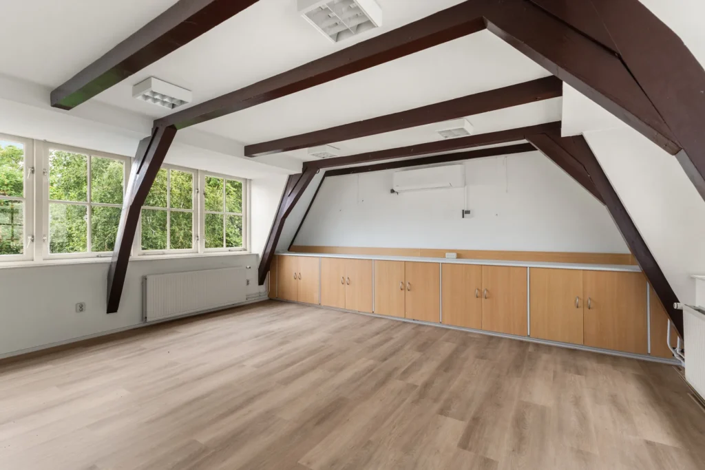 Lichte, lege kamer met houten vloer en grote ramen met zicht op groen.