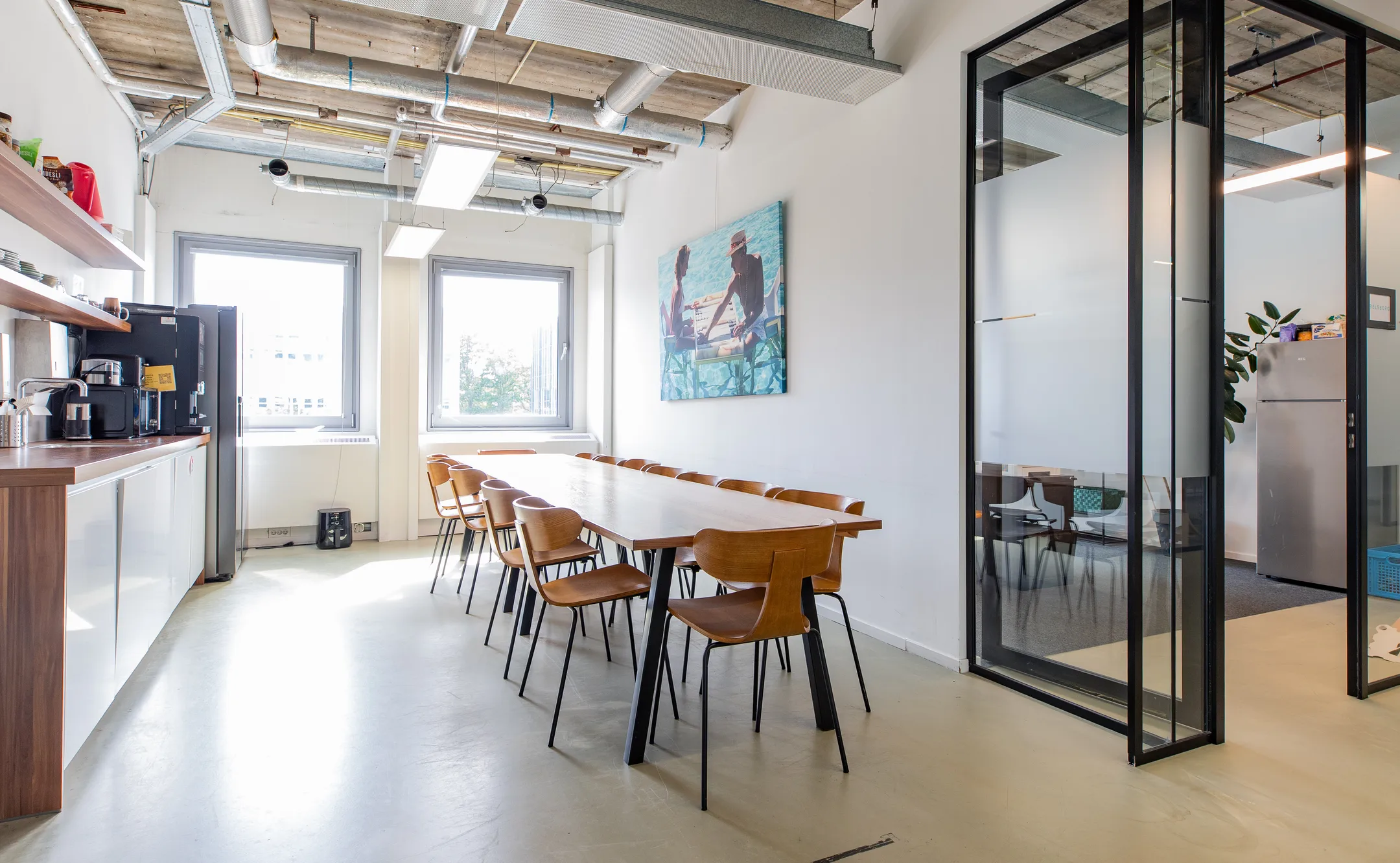 Een moderne kantoorruimte met een lange tafel omringd door houten stoelen en een keukenhoek met koffiezetapparaat.