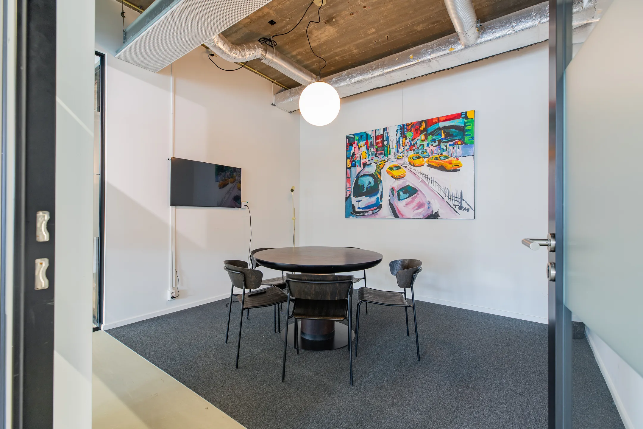 Een vergaderruimte met een ronde tafel en stoelen, een kleurrijk schilderij aan de muur en een televisie.
