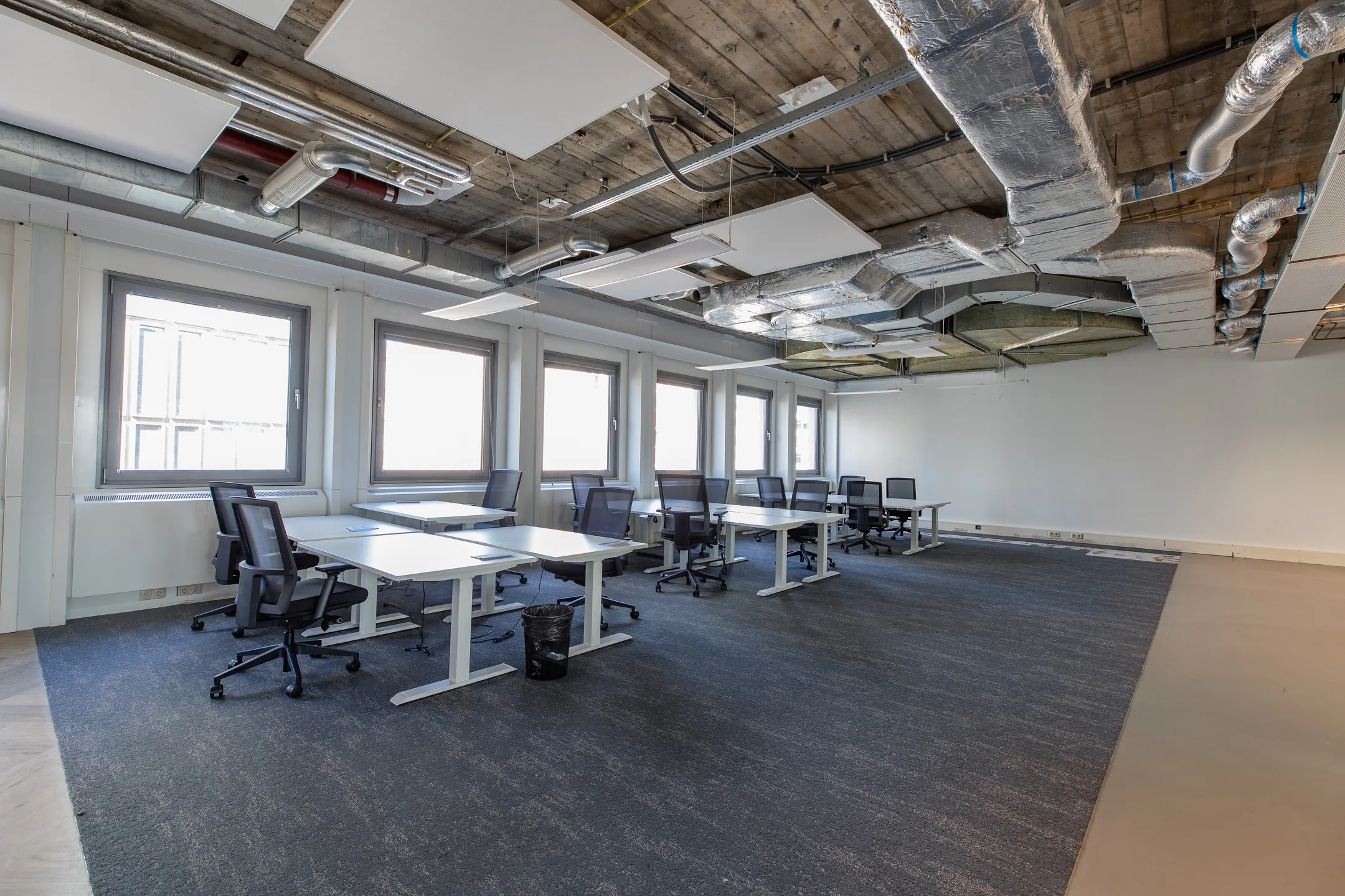 Een moderne, open kantoorruimte met bureaus en bureaustoelen, met zichtbare leidingen aan het plafond.