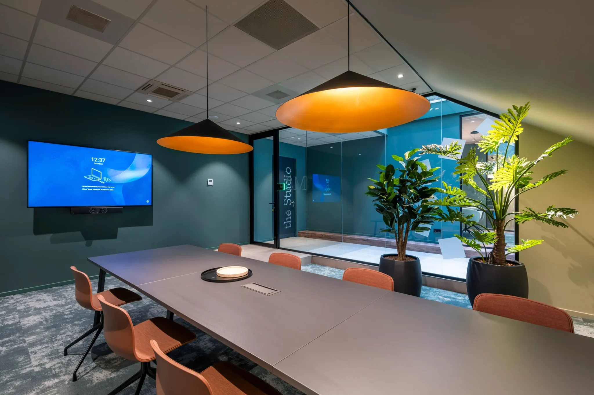 Moderne vergaderruimte genaamd "the Studio" met een lange tafel, designstoelen, grote hanglampen, een scherm aan de muur en kamerplanten bij een glazen wand.