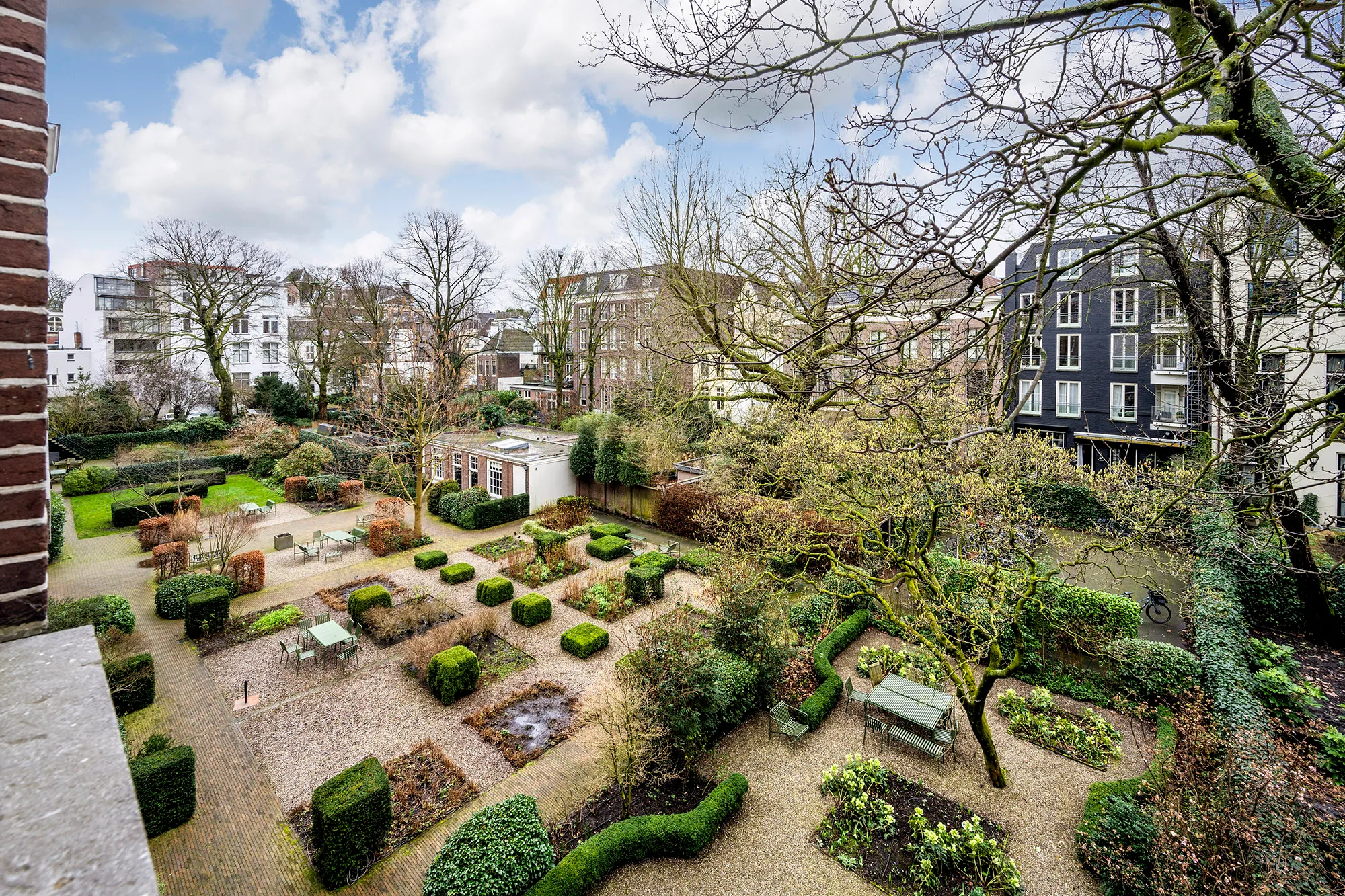 Een groene, goed verzorgde stadstuin met wandelpaden, struiken en bomen, omringd door gebouwen.