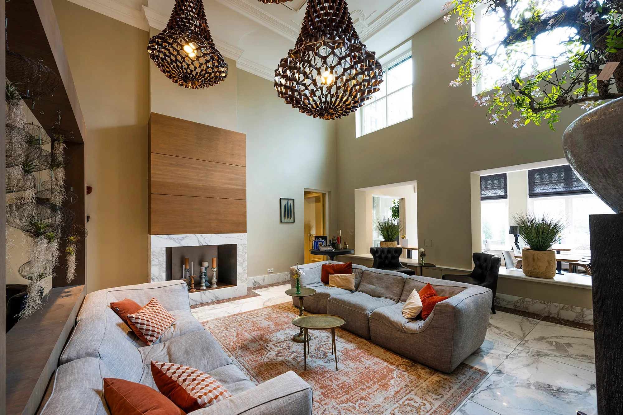 Een elegante woonkamer met grote ramen, moderne houten en marmeren accenten, grijze banken, warme kussens en decoratieve hanglampen.