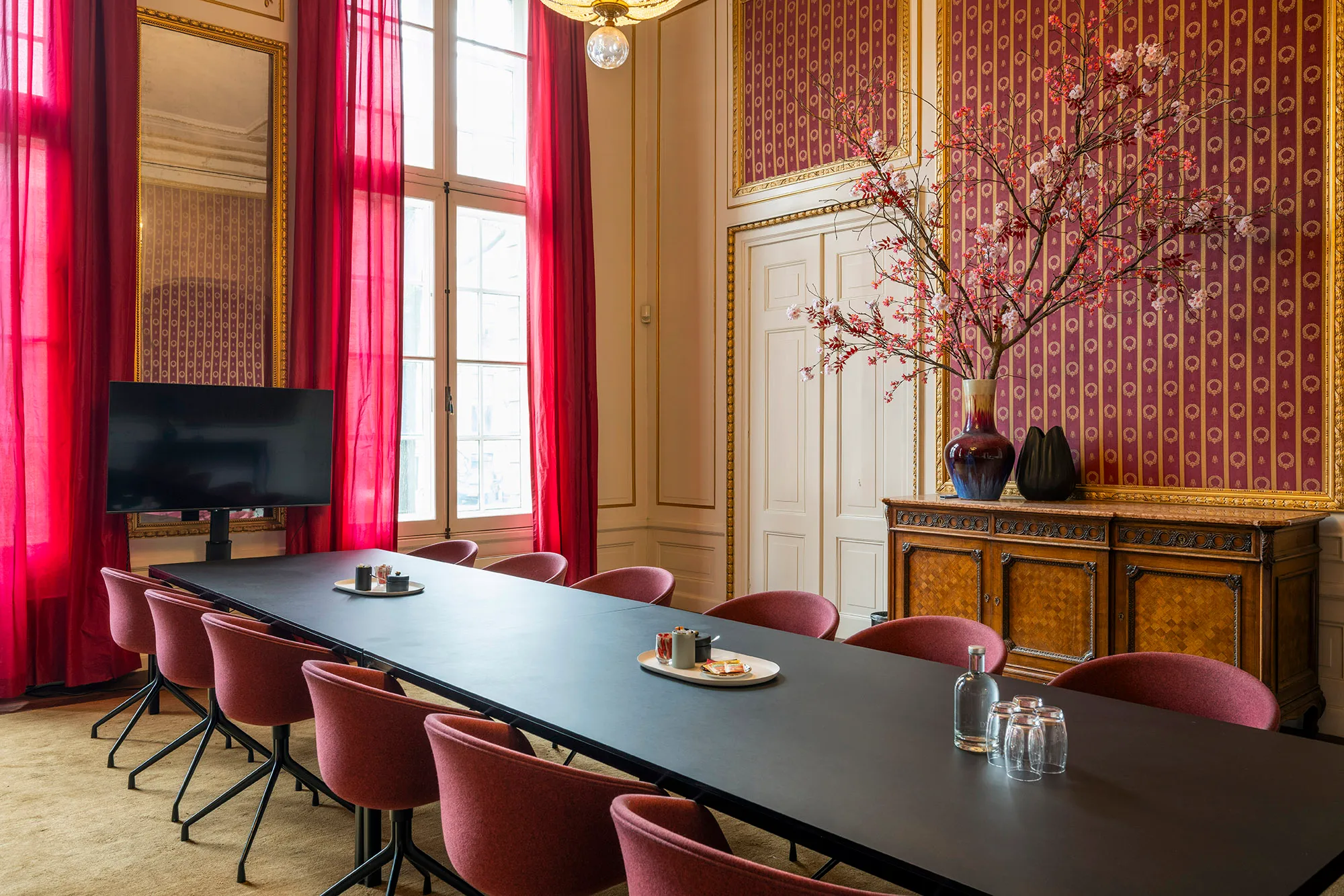 Een elegante vergaderruimte met een lange zwarte tafel, rode stoelen, een tv-scherm en een sierlijk bloemstuk op een kast.