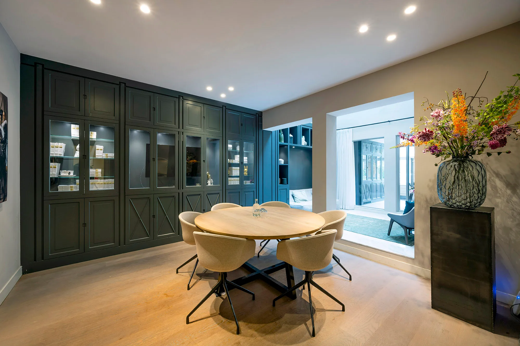 Een moderne eetkamer met een ronde tafel, beige stoelen, een donkere wandkast en een vaas met kleurrijke bloemen.