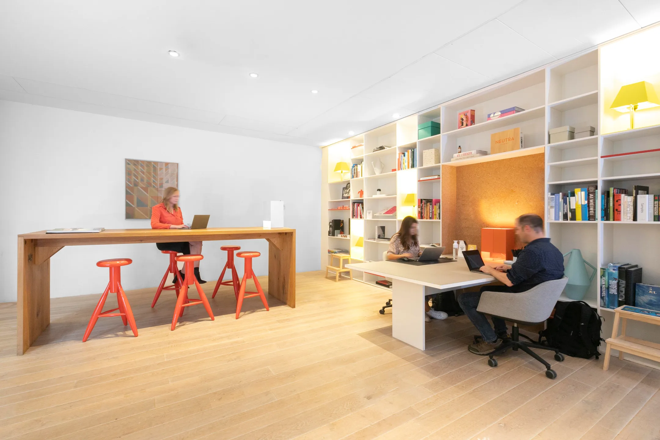 Een moderne kantoorruimte met witte muren, een grote houten tafel, rode krukken en mensen die op laptops werken naast een grote boekenkast.