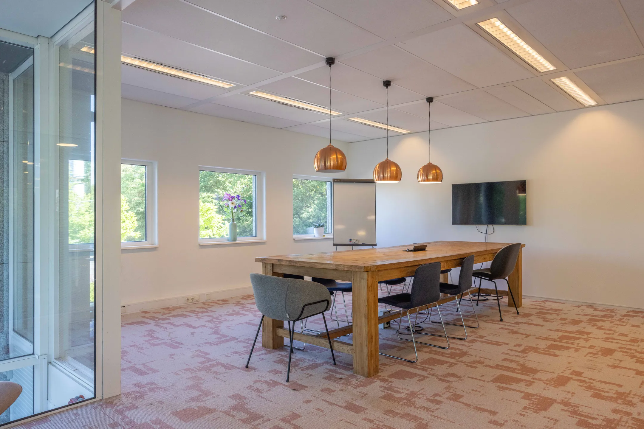 Een moderne vergaderruimte met een houten tafel, stoelen, grote ramen en hanglampen.