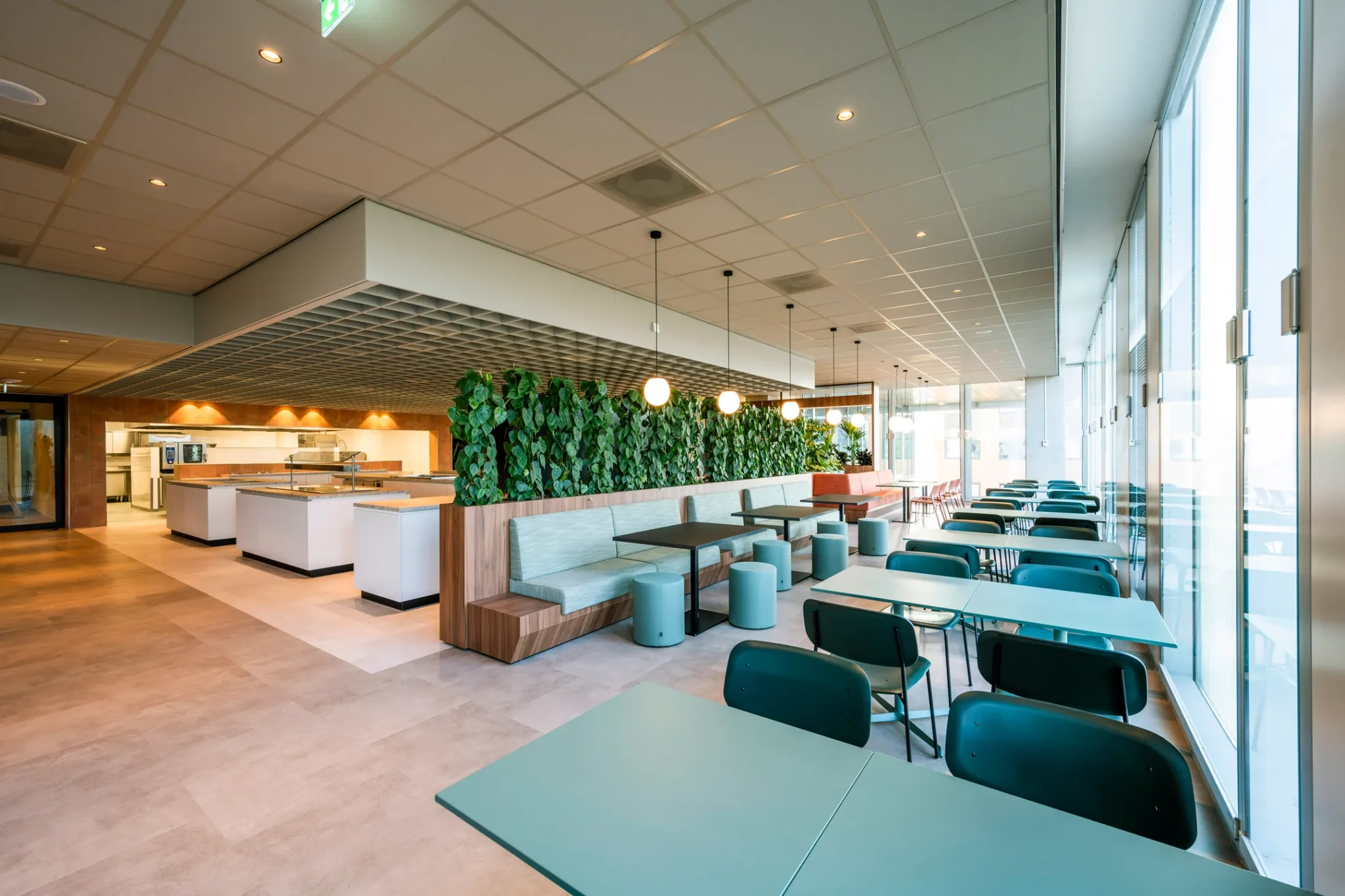 Moderne en ruim opgezette bedrijfs- of schoolkantine met licht interieur, groene accenten en zitplekken aan lange tafels en banken.