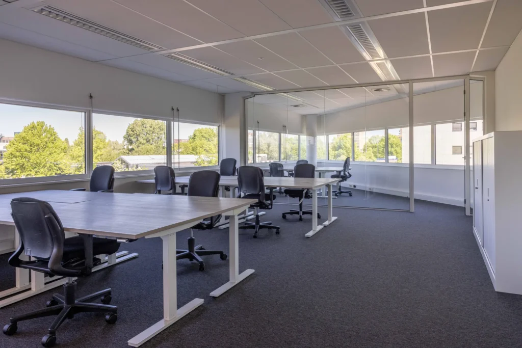 Een moderne, lichte kantoorruimte met bureaus en stoelen, omgeven door grote ramen.