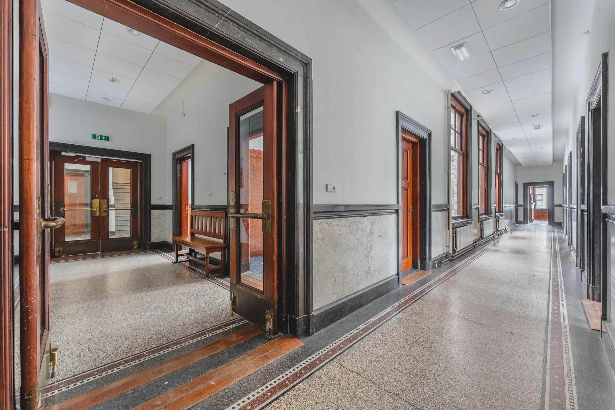 Een lange, lichte gang met houten deuren en een marmeren lambrisering in een historisch gebouw.