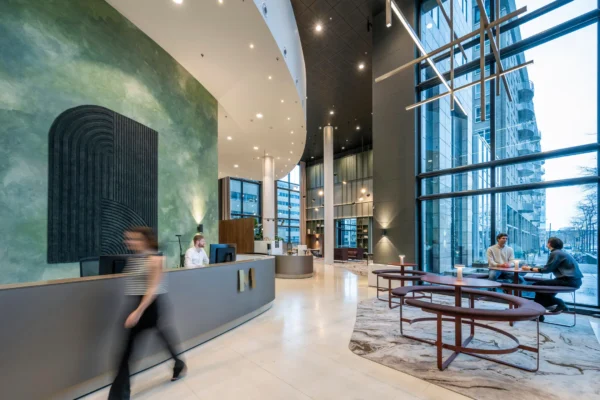 Een moderne hotellobby met een receptie, personen aan het werk en mensen in gesprek bij het raam.
