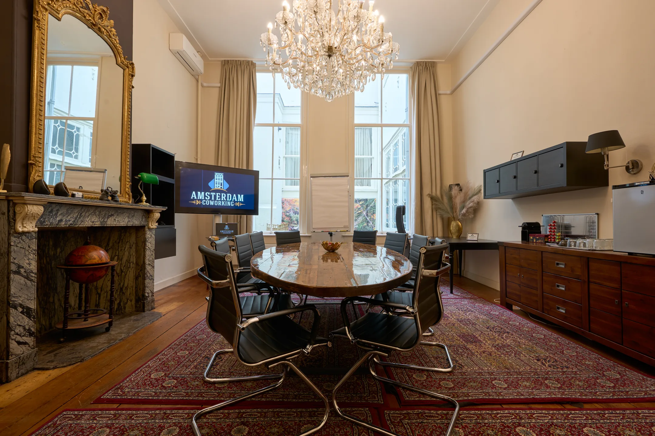 Vergaderruimte met een ovale tafel, stoelen, grote ramen en een kroonluchter, met op de achtergrond een tv-scherm met "Amsterdam Coworking".