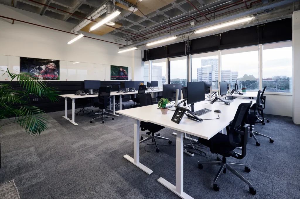Een modern en ruim kantoor met bureaus, computers en grote ramen met uitzicht op gebouwen.