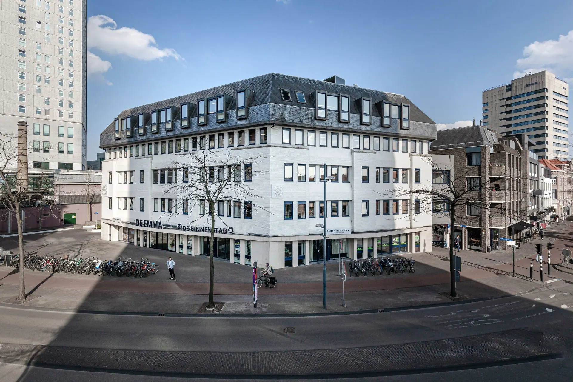 Wit kantoorgebouw De Emma aan een drukke straat in de binnenstad met fietsenrekken en enkele voorbijgangers.