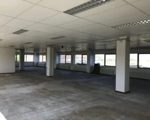 Lege kantoorruimte met systeemplafond, betonnen vloer en grote ramen rondom.