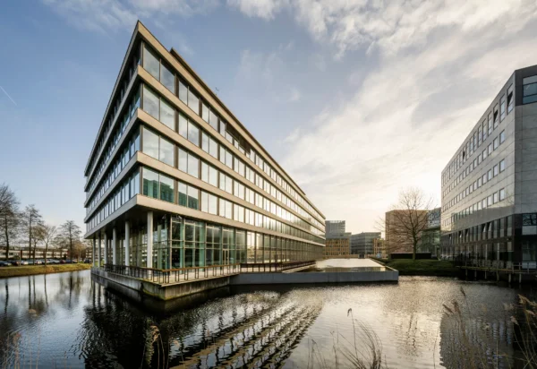 Een modern kantoorgebouw met grote ramen gelegen naast een waterpartij onder een helderblauwe lucht.