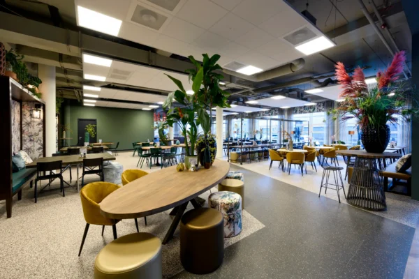Een moderne kantine met kleurrijke stoelen en tafels, versierd met grote planten in vazen.