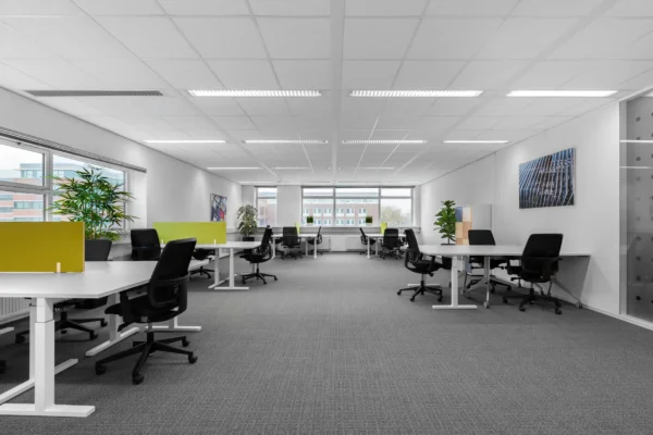 Een ruime, moderne kantoorruimte met bureaus, zwarte stoelen en groene scheidingspanelen.