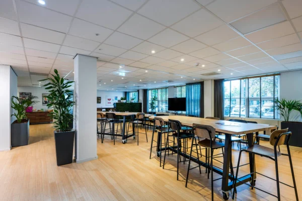 Een moderne, ruime vergaderruimte met hoge tafels, stoelen en grote ramen, gevuld met veel natuurlijk licht en groene planten.