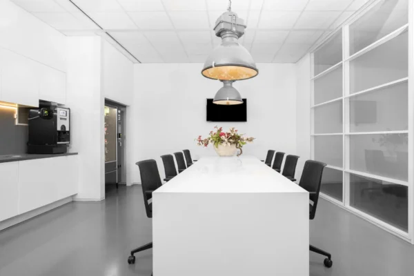 Een moderne vergaderruimte met een lange witte tafel, omringd door zwarte stoelen en verlicht door grote hanglampen.